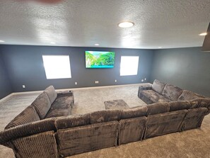 A living room with a large sectional sofa, two windows, and a flat-screen TV mounted on the wall.