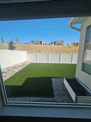A view through a window of a backyard with artificial grass, a white fence, and a hillside with houses in the background.