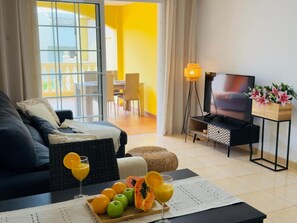 Ein modernes Wohnzimmer mit einer Couch, einem Couchtisch mit Früchten, einem Fernseher und einem Balkon mit gelben Wänden.