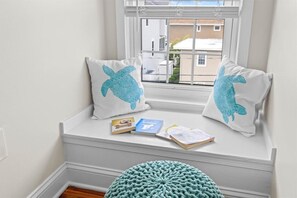 En sittplats vid fönstret med två kuddar med ett motiv av havssköldpaddor, en stickad pall och utsikt över byggnaderna utanför.