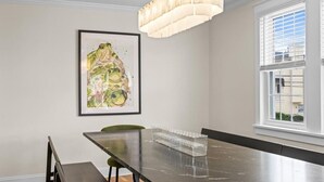 Un comedor con una larga mesa, sillas, una araña, una obra de arte enmarcada y una ventana con persianas.
