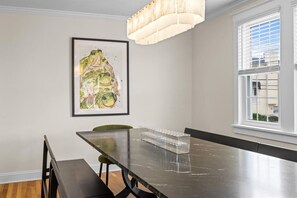 A dining area with a long table, chairs, a chandelier, a framed artwork, and a window with blinds. - Just Renovated Coastal Retreat | Outdoor Lounge (Ocean City)
