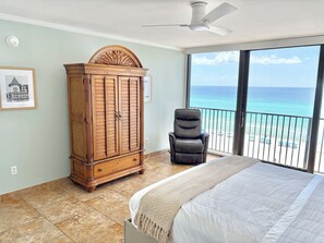 Une chambre à coucher avec un lit, une armoire en bois, une chaise noire, un ventilateur de plafond et un balcon avec vue sur l’océan.