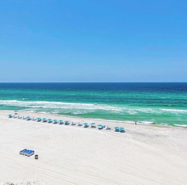 Ein Strand mit einer Reihe von Sonnenschirmen und Liegestühlen, einem klaren Himmel und dem Meer im Hintergrund.