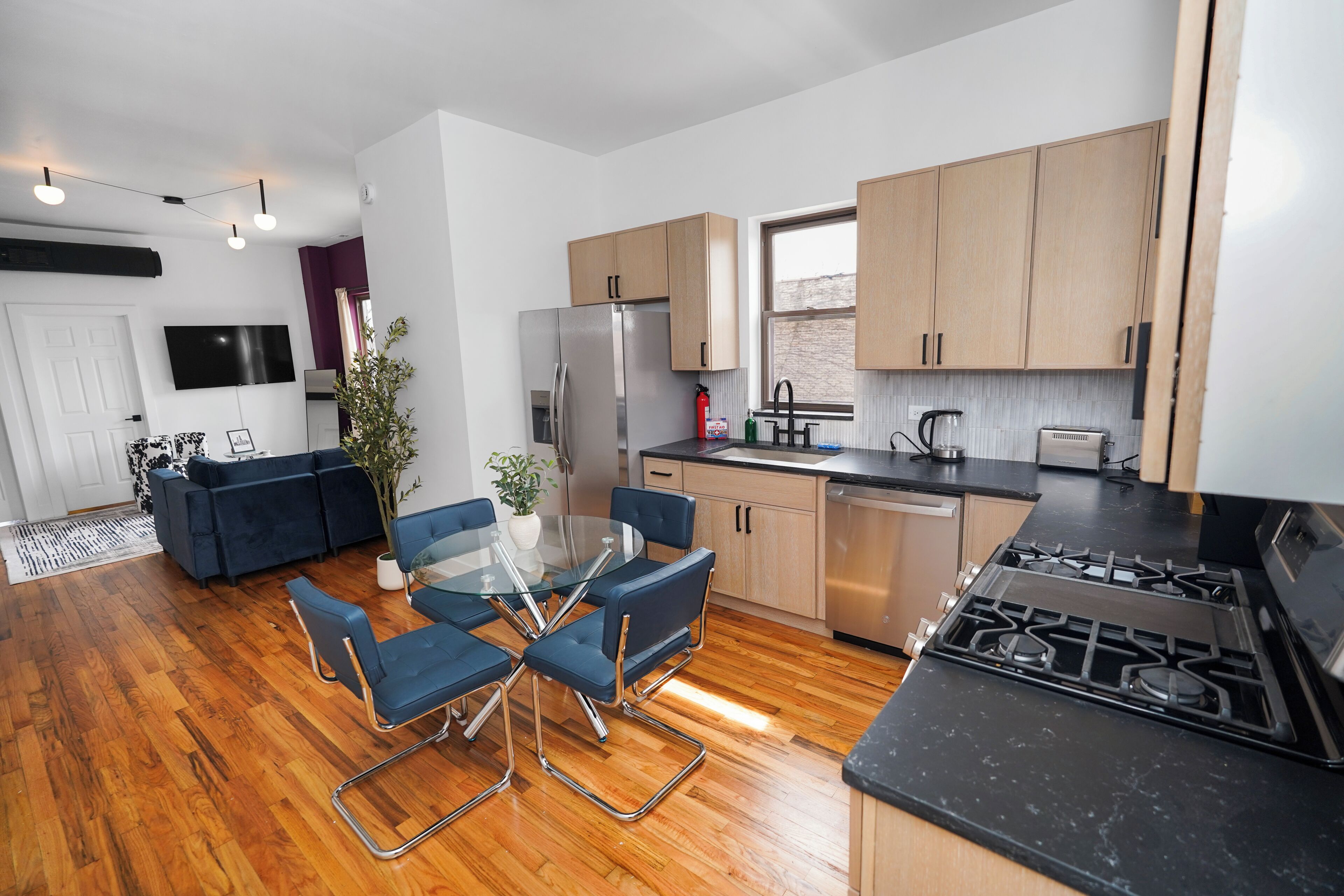 A modern kitchen with a dining area, wooden cabinets, a stainless steel refrigerator, and a gas stove.