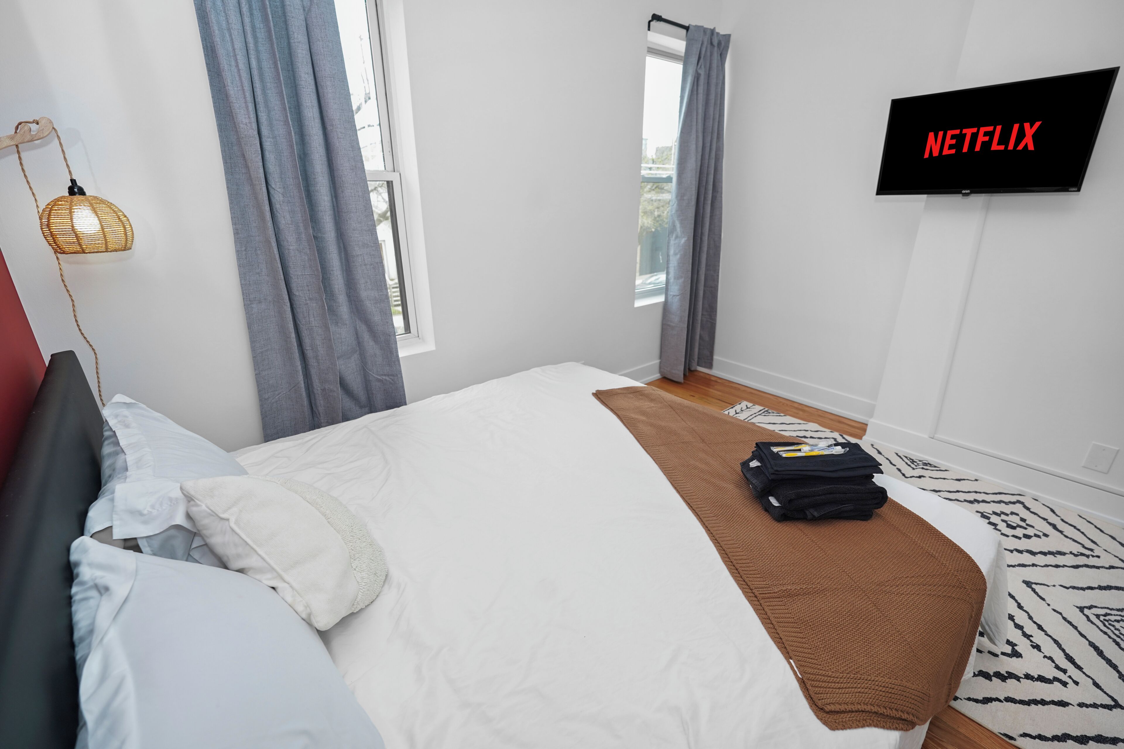 A modern hotel room with a bed, a TV mounted on the wall, a window with curtains, and a woven light fixture.