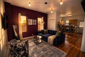 A living room with a sofa, coffee table, and patterned rug. There is a dining area with chairs and a table. The room has a large window with curtains, and the walls are painted in a dark color. - Modern 3BR Chicago Retreat, 7 Min to Fulton Market (Chicago)