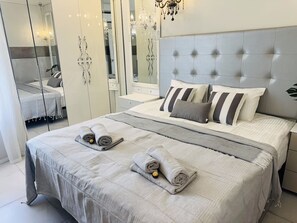 Ein ordentlich bezogenes Bett mit grauer Decke, Kissen und aufgerollten Handtüchern.