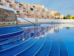 Ein Schwimmbecken mit geometrischem Fliesenmuster, einer Steinwand und Spiegelungen von Gebäuden im Wasser.