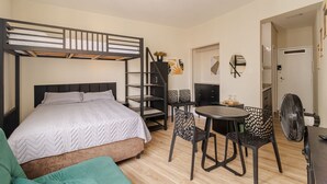 Una cama individual superpuesta con escalera, un escritorio con sillas, un ventilador y una puerta que da a otra habitación.