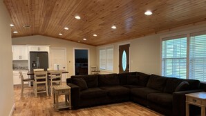 Ein geräumiger Wohnbereich mit Holzbalkendecke, einem dunkelgrauen Ecksofa, einem Esstisch mit Stühlen und einer Küche im Hintergrund.