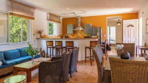 Una cocina moderna con una isla central, taburetes de bar y una zona de comedor con muebles de mimbre.