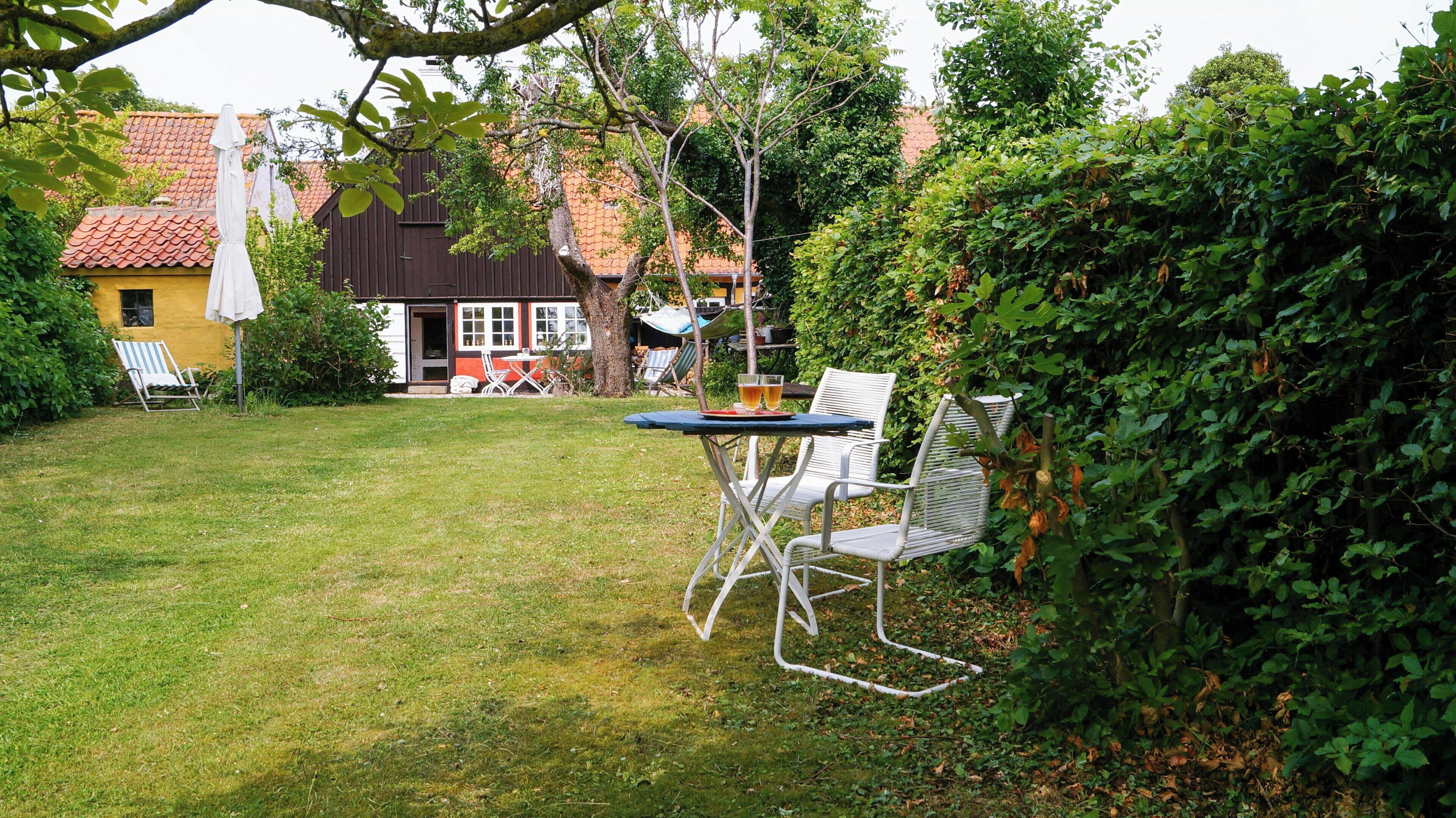 A grassy outdoor area with a table and chairs, a hammock, and a building with a red-tiled roof in the background.