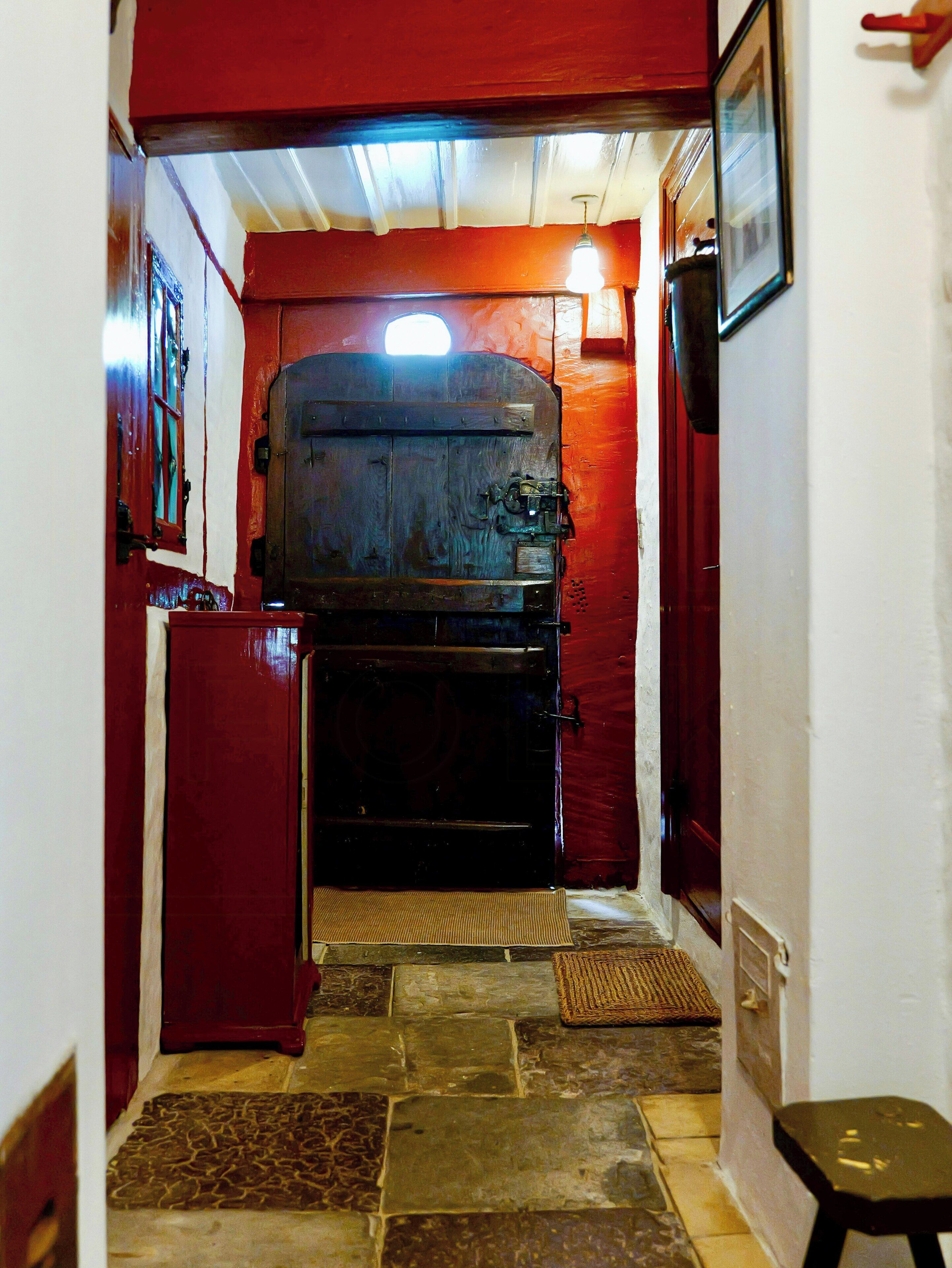 A narrow hallway with a wooden door at the end, a red wall, and a stone floor.