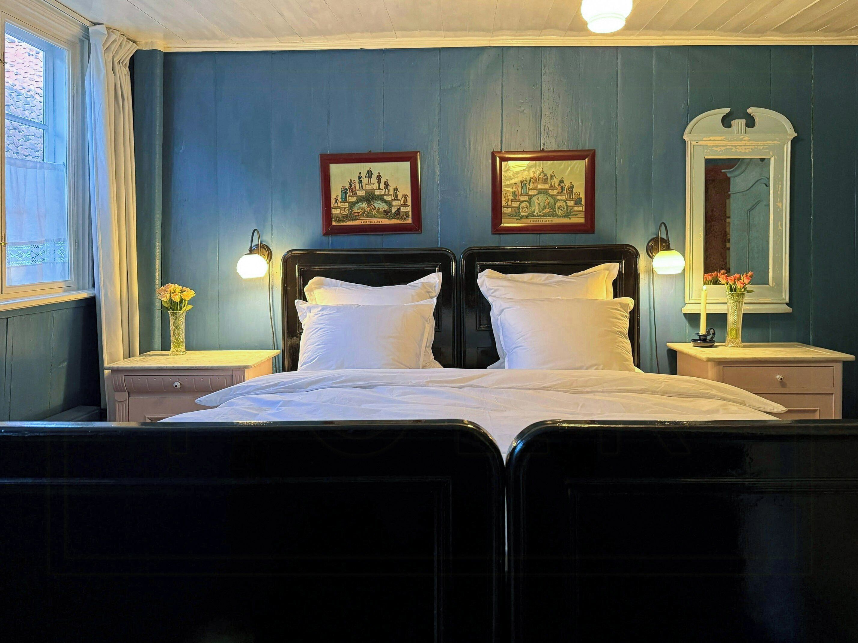 A double bed with white linens, two bedside tables with lamps, a mirror, and two framed pictures on the wall.