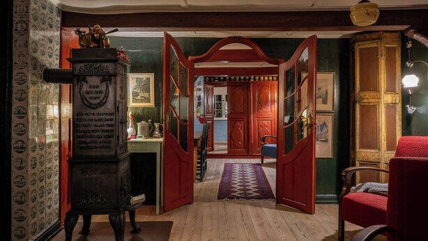 A room with a wooden floor, a red chair, a wooden cabinet, and a door leading to another room. - Historic stay in the heart of Aeroskobing (Ærøskøbing)