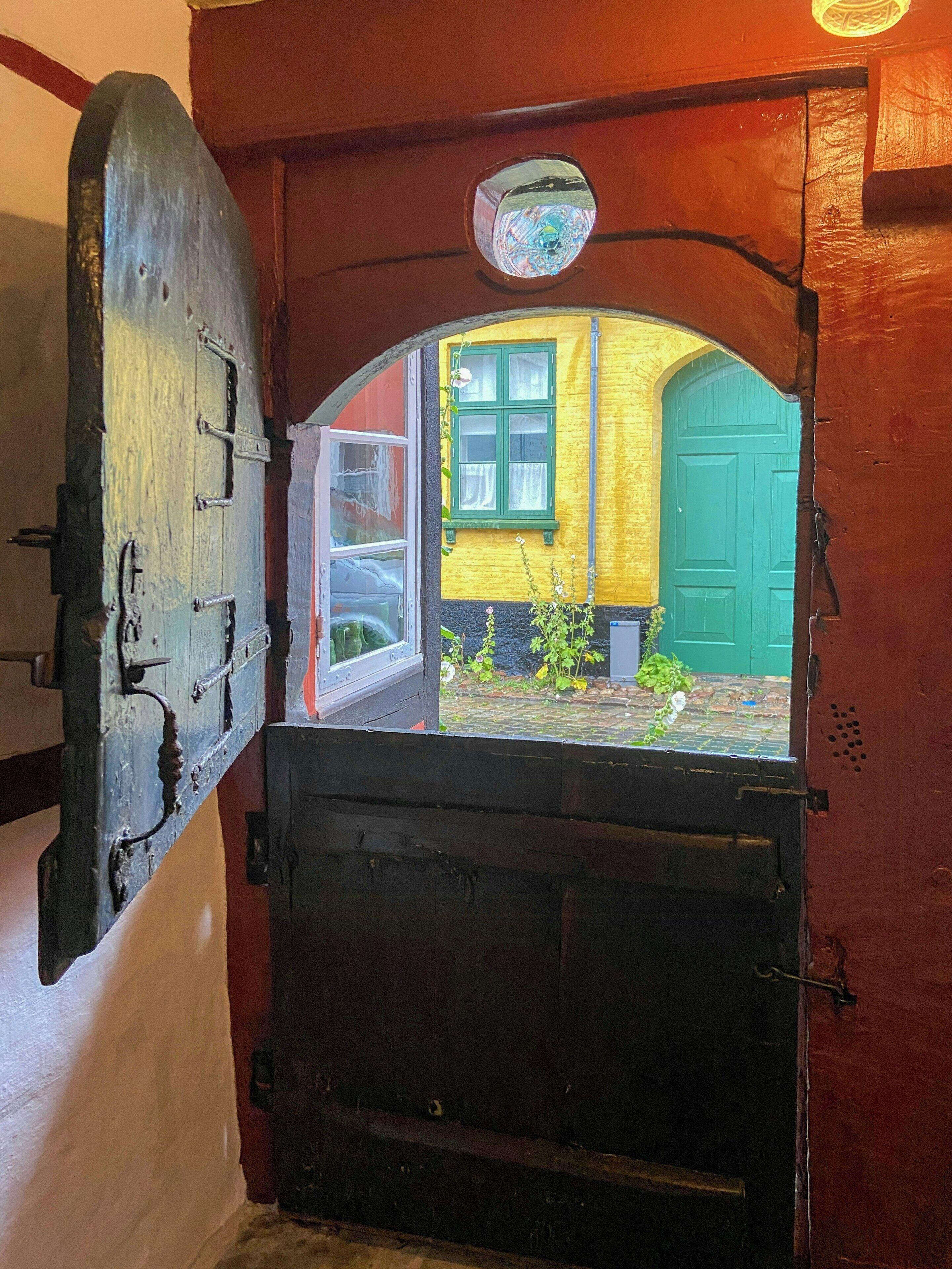 An open wooden door with a view of a yellow building and a green door.