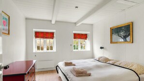 Um quarto com uma cama, duas janelas com cortinas vermelhas, uma cômoda vermelha e um quadro na parede.