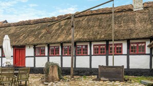 Um edifício tradicional com telhado de palha, paredes brancas com estrutura de madeira escura, janelas vermelhas e um pátio pavimentado em pedra.