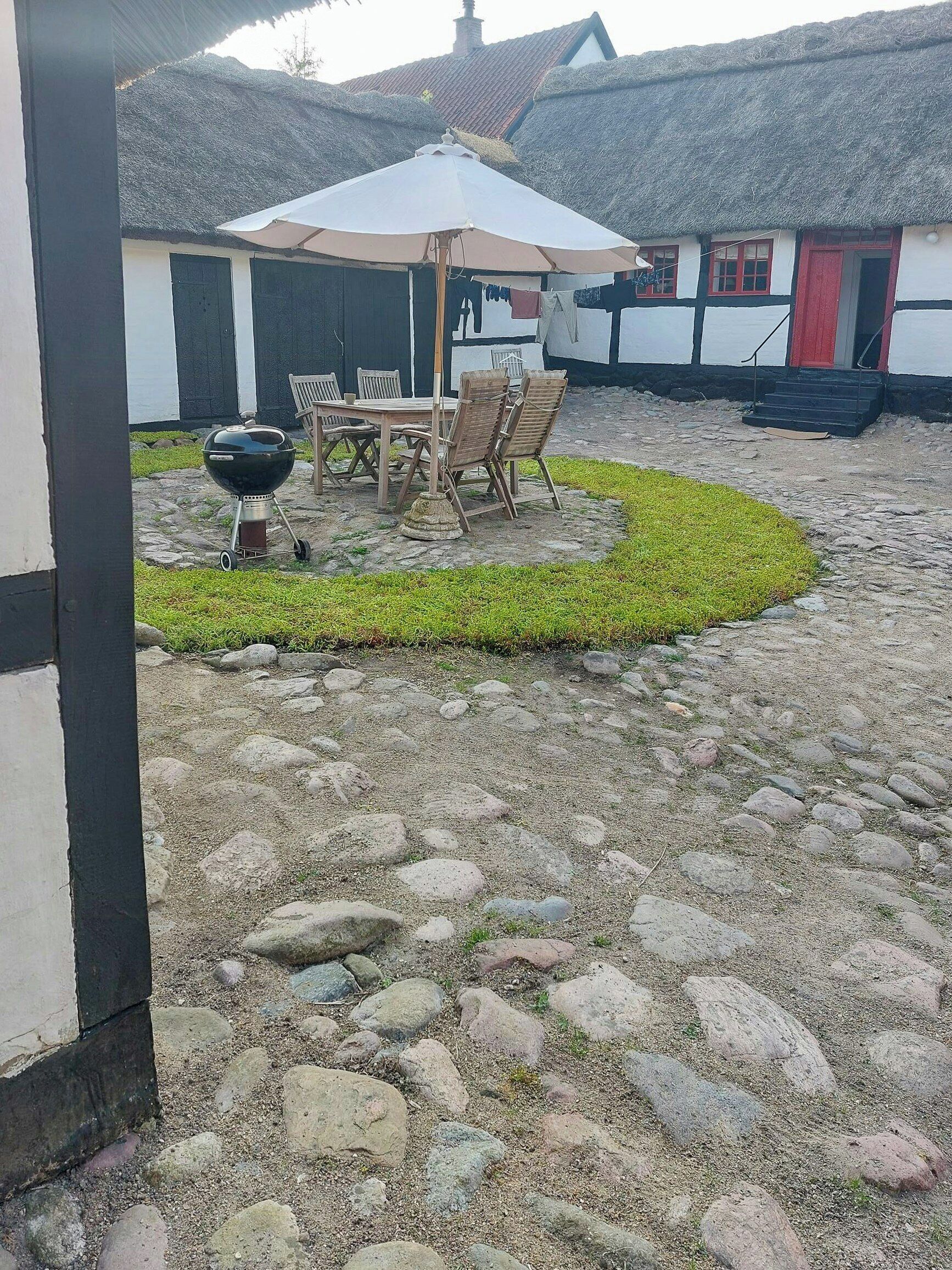 En terrasse med et bord og stole, en grill og en bindingsværksbygning i baggrunden.