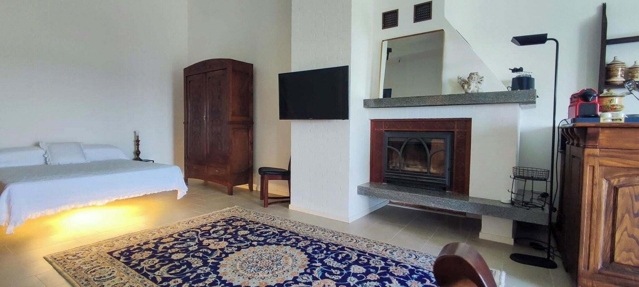 A bedroom with a bed, a wooden wardrobe, a television, a fireplace, and a rug.