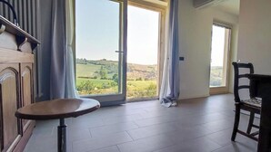 Een kamer met een groot raam, een houten stoel, een klein rond tafeltje en uitzicht op een groen landschap.