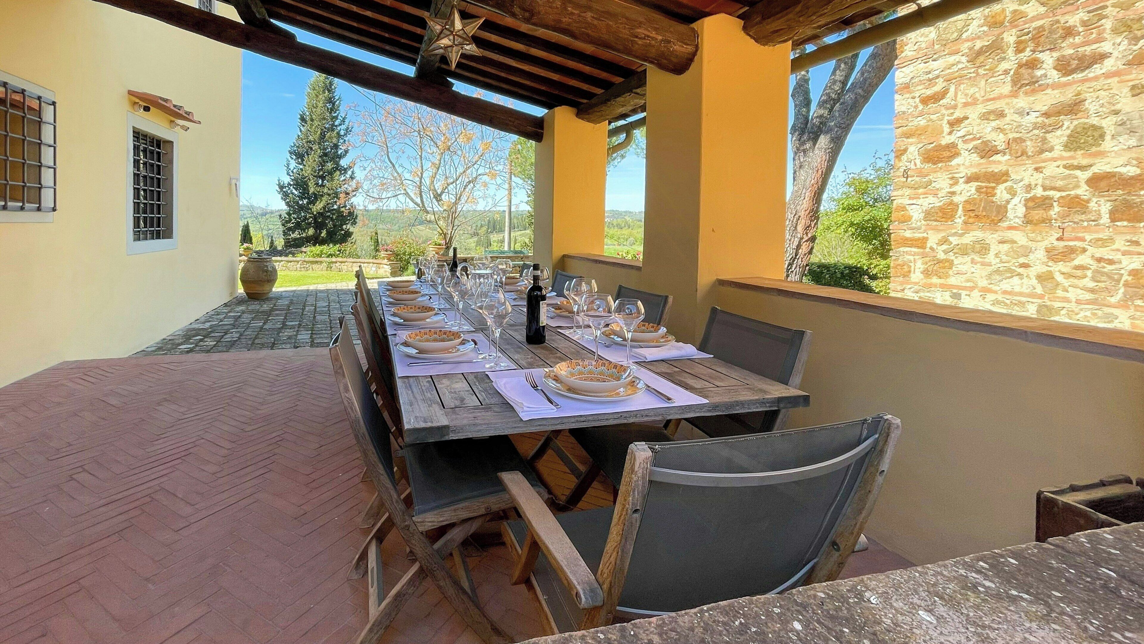 Un área de comedor al aire libre con una larga mesa de madera dispuesta para una comida, rodeada de muros de piedra y vistas al campo.