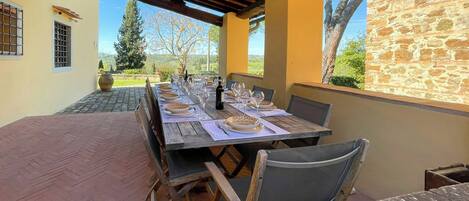 Un área de comedor al aire libre con una larga mesa de madera dispuesta para una comida, rodeada de muros de piedra y vistas al campo.