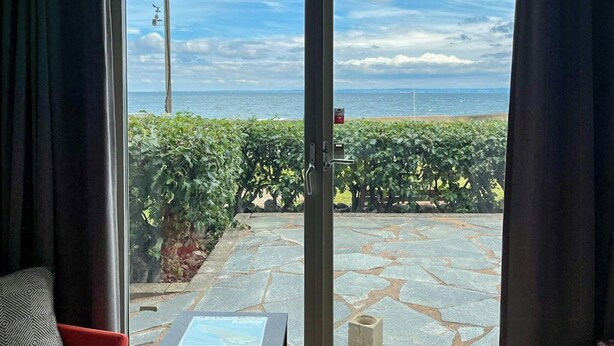 Une pièce dotée d’une porte-fenêtre coulissante donnant sur un patio avec un sol en pierre, une vue sur la mer et une haie.