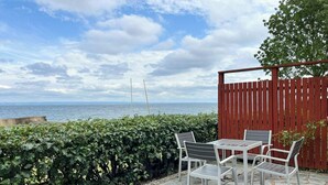 Une terrasse avec un sol en pierre, une table et des chaises, donnant sur une étendue d’eau et un ciel nuageux.