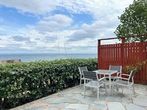 Eine Terrasse mit Steinbelag, einem Tisch und Stühlen, mit Blick auf ein Gewässer und einen bewölkten Himmel.