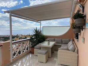 Ein Balkon mit Korbmöbeln, einem Glastisch und Topfpflanzen.