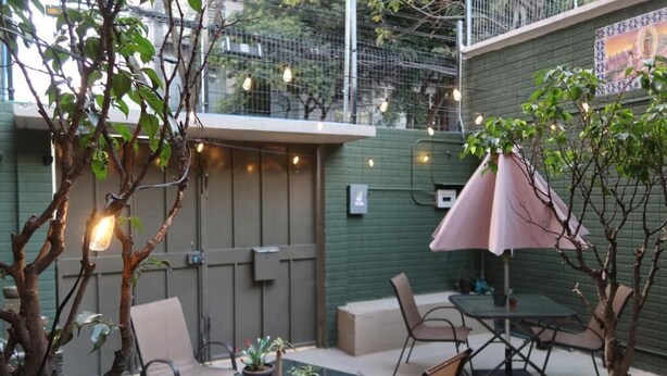 Un patio al aire libre con mesas y sillas, una pequeña mesa con plantas en macetas y luces colgantes.