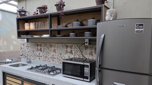 Cocina compacta con estufa a gas, microondas y refrigerador, con repisas superiores que albergan utensilios de cocina y plantas en macetas.