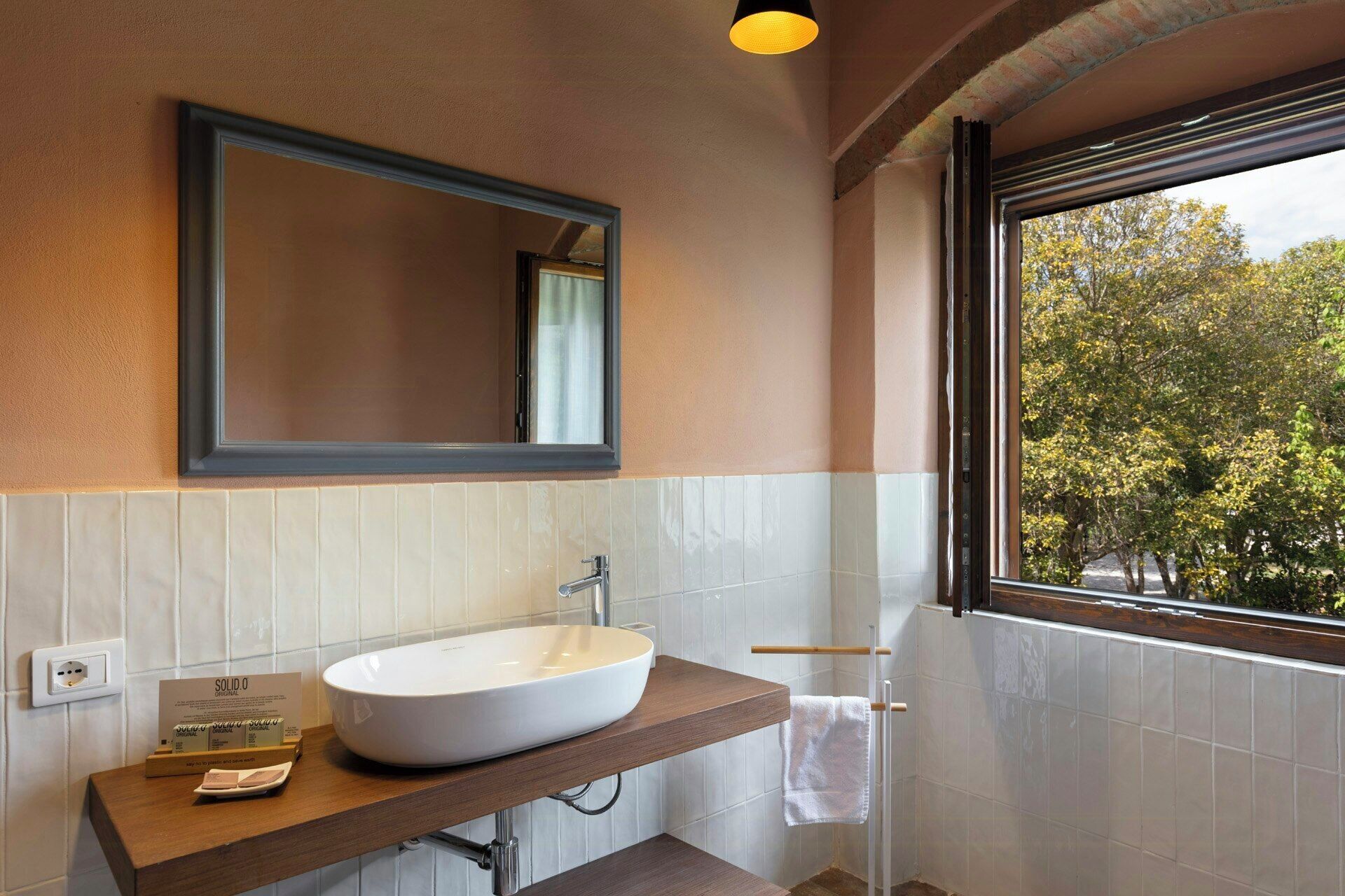 Ein Badezimmer mit einem weißen Waschbecken, einem Spiegel und einem Fenster mit Blick auf Bäume.