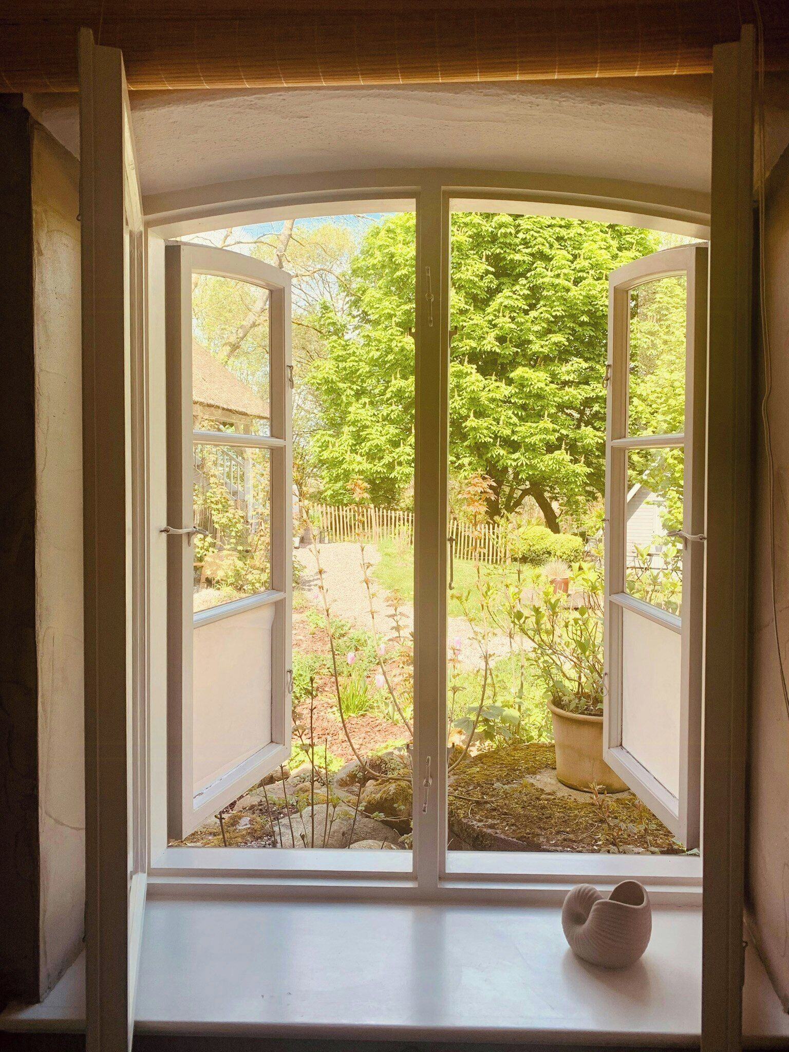 A bay window with an open shutter, a white vase on the sill, and a view of a garden with trees and a wooden fence.