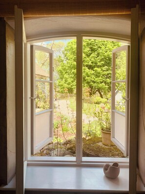 Ein Erker mit geöffnetem Klappfenster, eine weiße Vase auf der Fensterbank und Blick auf einen Garten mit Bäumen und einem Holzzaun.