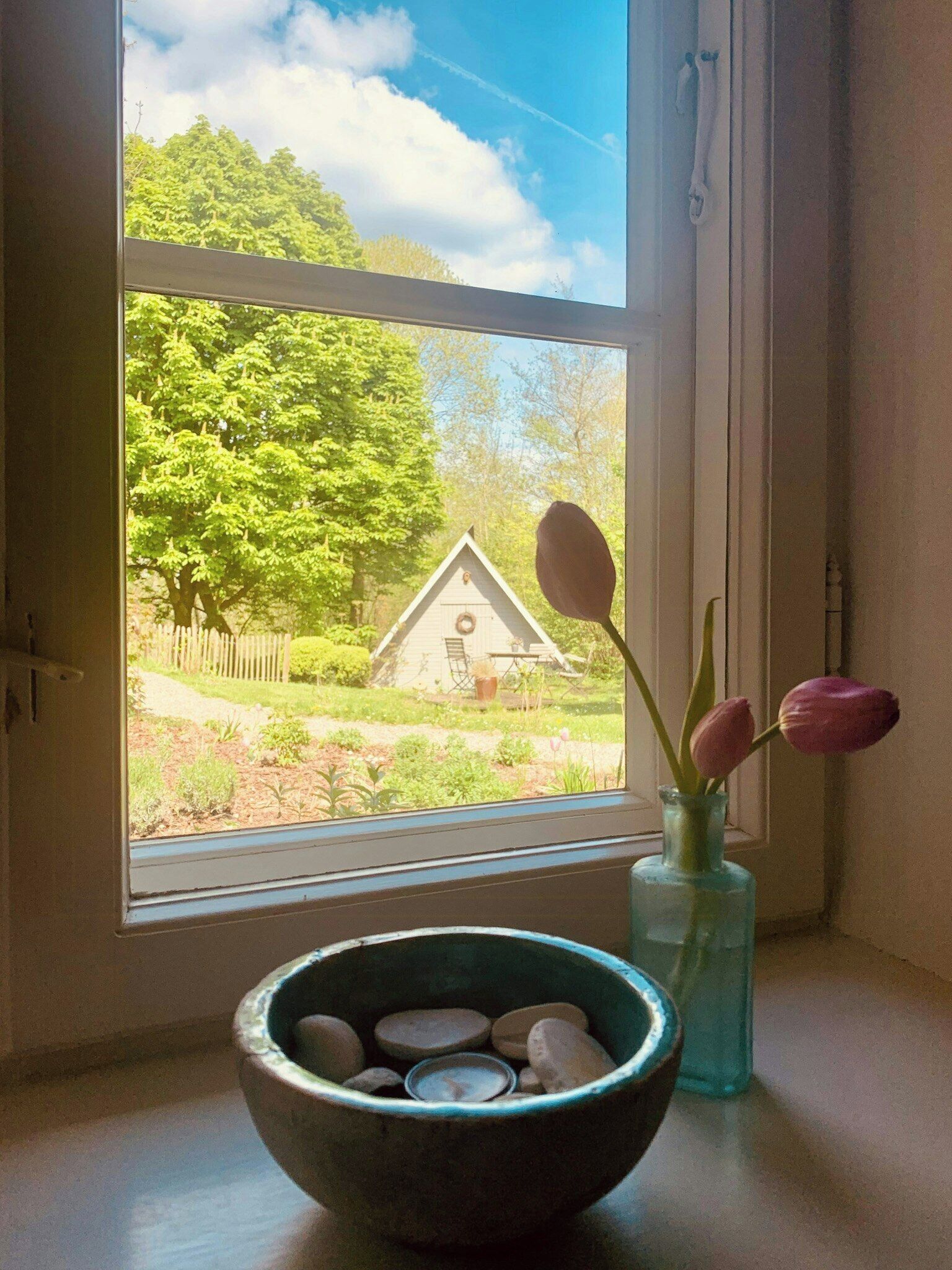 A window with a view of a house, a bowl with stones, and a vase with flowers.