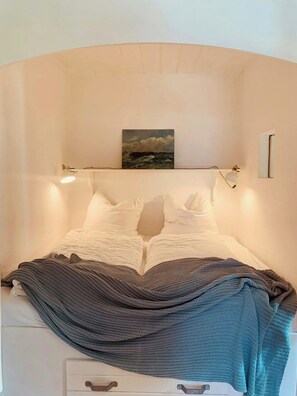 Ein ordentlich bezogenes Bett mit weißen Bettwäsche, einer dunkel gestrickten Decke und einem Gemälde darüber.
