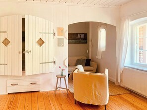 Ein Zimmer mit Holzboden, einer weißen Tür mit einem rautenförmigen Fenster, einem beigefarbenen Sofa und einem kleinen Hocker mit Blumen in einer Vase.