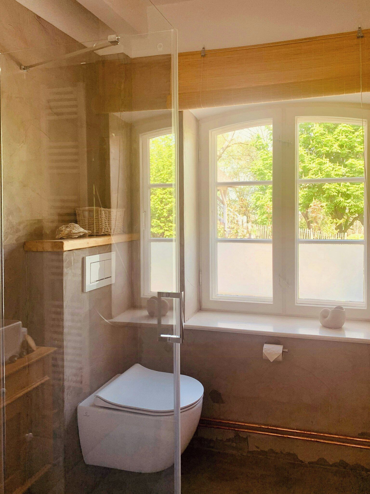 Un baño con cabina de ducha de vidrio, inodoro, ventana con vista a árboles y una pequeña repisa con una cesta.