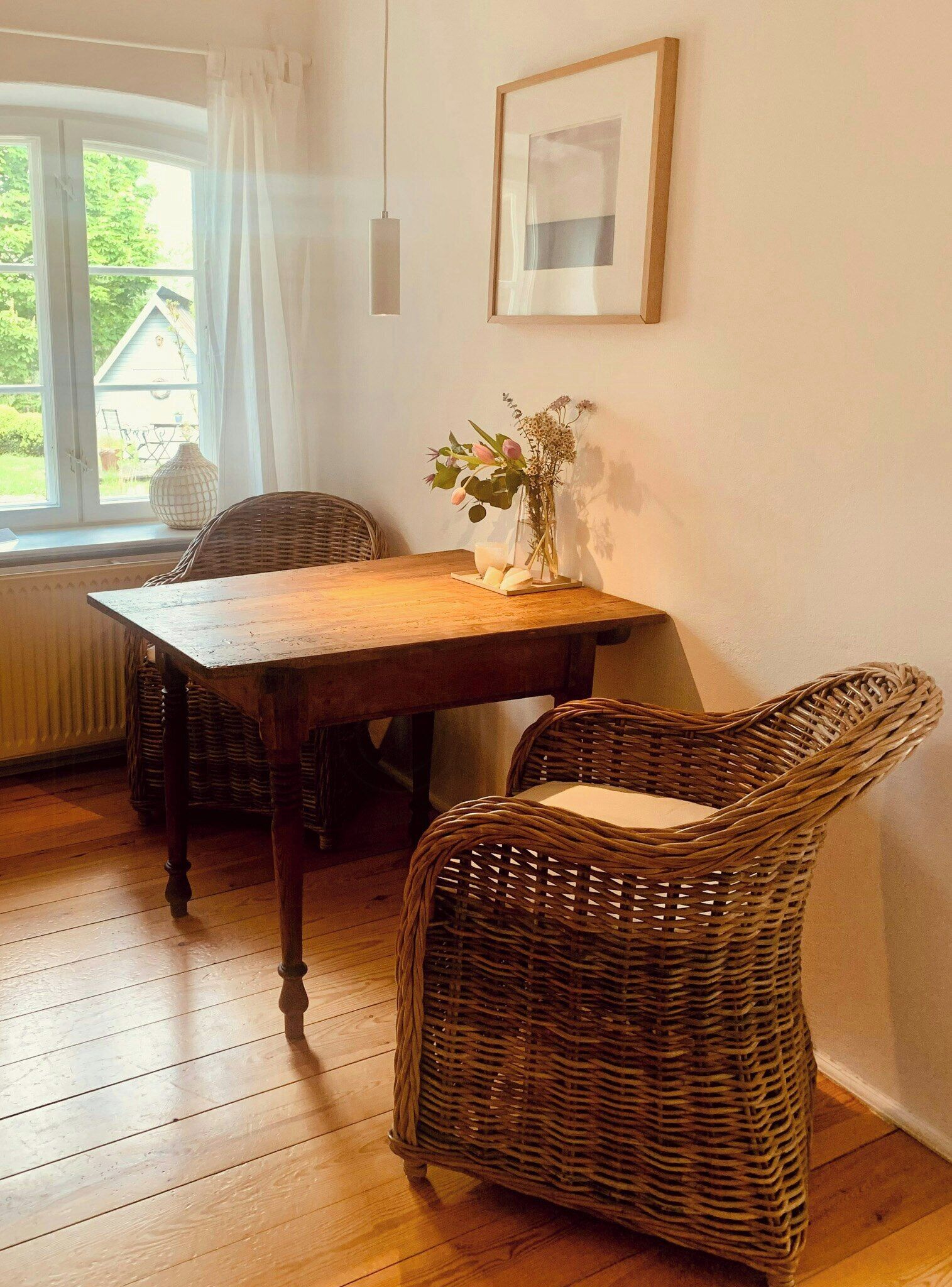 A dining area with a wooden table, wicker chairs, a vase with flowers, a framed picture on the wall, and a window with curtains.