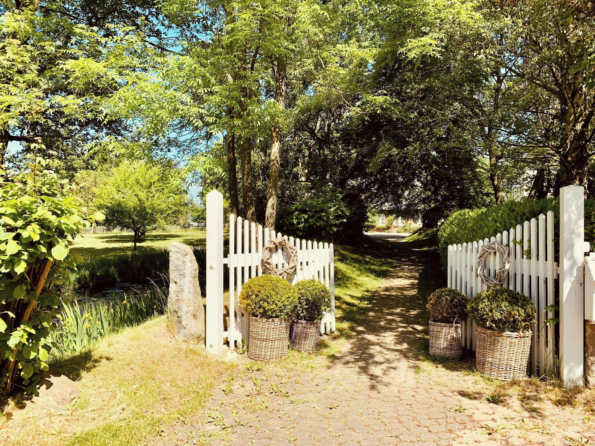 A pathway leading through a garden with a white picket fence, stone pillars, and potted plants.