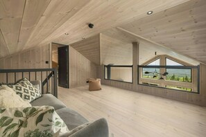 A spacious loft bedroom with a wooden ceiling, a large window offering a view of the outdoors, and a cozy seating area with a sofa and cushions.