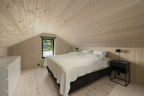 Une chambre avec un plafond en pente, un lit avec une literie blanche, une table de chevet avec une lampe et une fenêtre donnant sur un paysage verdoyant.