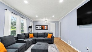 A living room with a sectional sofa, ottoman, and a flat-screen TV mounted on the wall. - Luxury Metro Atlanta Retreat - Sleeps 10 (Decatur)
