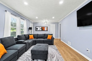 Ein Wohnzimmer mit Ecksofa, Hocker und einem Wand-Fernseher.