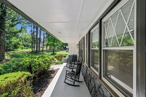 Eine überdachte Veranda mit drei Schaukelstühlen, einer Steinmauer und Blick auf einen gepflegten Garten und Bäume.
