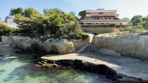 A coastal area with a stone bridge leading to a building with a modern design, surrounded by greenery and clear water. - House with view of Ortigia and sea access (Siracusa)
