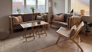 A living room with a sofa, a coffee table, and a chair. There are windows with a view of buildings and a plant on the windowsill.